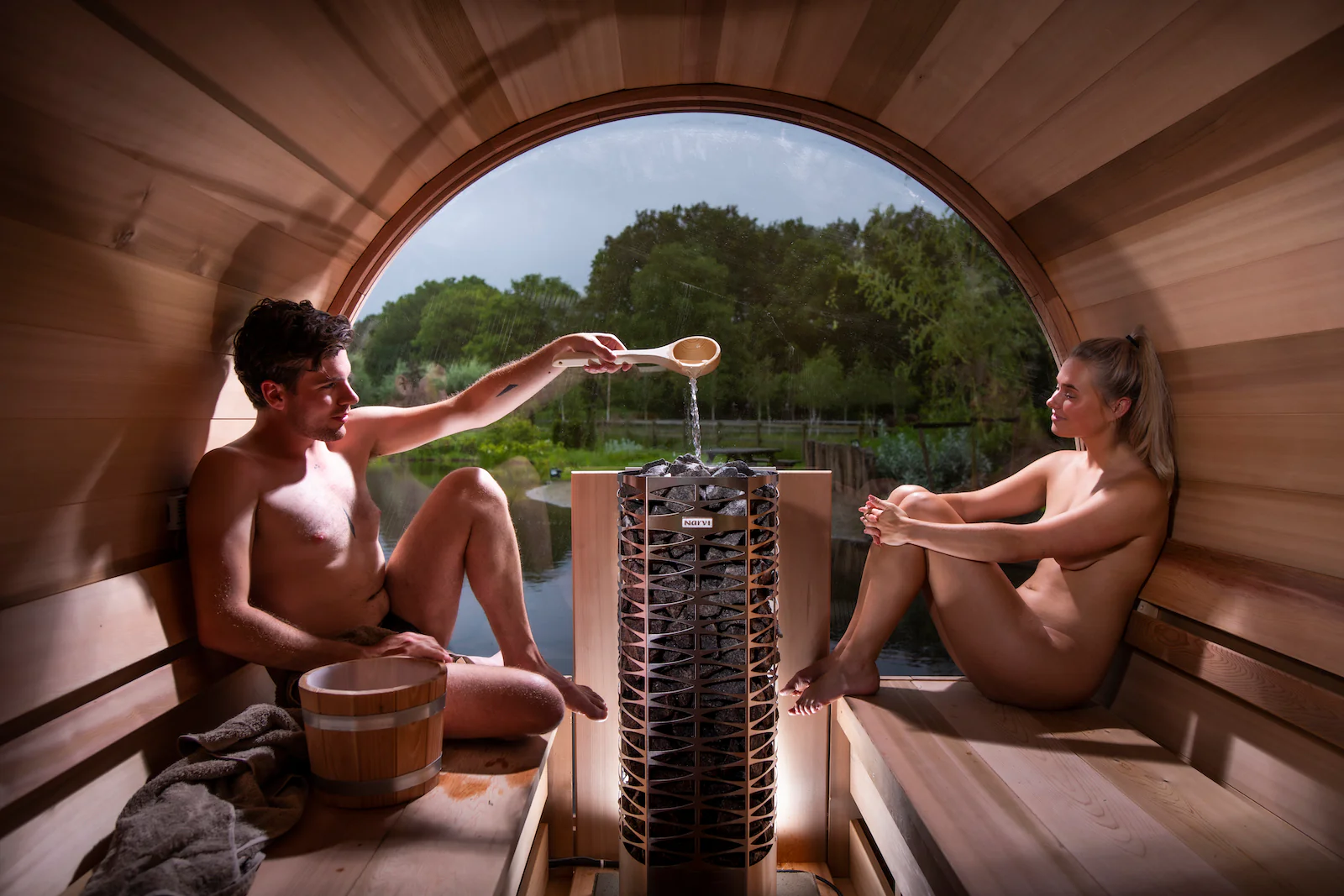 Een jong stel aan het genieten in een barrelsauna met een elektrische kachel in het midden met een prachtig uitzicht over een meer